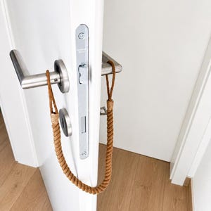May include: A brown twisted rope door handle accessory featuring two loops attached to a silver metal door lever. The rope is secured with a wrapped cord detail at the top of each loop, creating a decorative pull for the door handle mechanism.