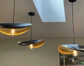 May include: Three gold and black pendant lights hang from the ceiling. The lights are shaped like shallow bowls with a textured surface. The lights are illuminated with warm white light bulbs.