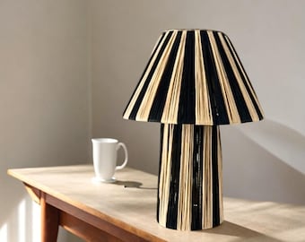 Puede incluir: Una lámpara de mesa a rayas negras y beige con una base cilíndrica y una pantalla ancha y acampanada. La lámpara está sobre una mesa de madera.