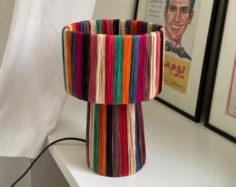 Könnte beinhalten: Eine bunte Tischlampe mit einem gestreiften Sockel und einem Schirm aus Garn. Die Lampe steht auf einer weißen Oberfläche mit einem gerahmten Poster dahinter.