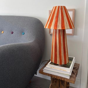 May include: A striped orange and white lamp with a woven shade sits on a wooden table with books stacked underneath. The lamp is next to a gray sofa.