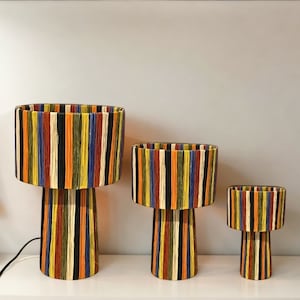 Può includere: Tre lampade da tavolo a righe colorate con una base cilindrica. Le lampade sono realizzate in materiale intrecciato e hanno un design moderno e unico.