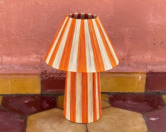 May include: A striped lamp with an orange and white design. The lamp is made of woven material and has a cylindrical base. The lamp is sitting on a tiled floor.