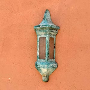 May include: A weathered, teal-colored outdoor lantern with a decorative top and bottom, mounted on a textured, orange-colored wall. The lantern has two rectangular glass panels. The lantern's aged appearance suggests it is made of metal.