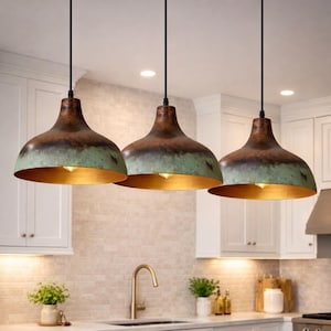 Pode incluir: Três luminárias pendentes com exterior cor de cobre e pátina verde. O interior de cada luminária é dourado. As luminárias pendem sobre uma ilha de cozinha com bancada de mármore. A cozinha tem armários brancos e um backsplash de azulejos neutros.