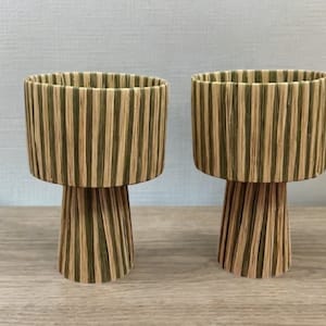 May include: Two wooden candle holders with a striped design in shades of brown and green. The holders have a wide, cylindrical top and a narrow, tapered base.