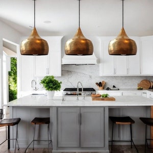 May include: Three gold pendant lights hang over a kitchen island with white cabinets and a gray countertop. The lights are teardrop shaped and have a hammered metal finish.