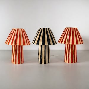 May include: Three table lamps with striped shades. The shades are made of woven paper and are in red, black, and orange. The lamps are on a white surface.