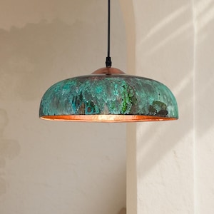 Op de afbeelding: Een hanglamp van koper met een verdigris afwerking. De koepelvormige kap heeft een textuur met groen- en blauwtinten. De lamp wordt opgehangen aan een zwart snoer. Diameter: ongeveer 30 cm.