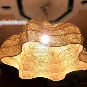 May include: A decorative hanging light fitting with a unique, layered design. The light fitting is made of a light brown, textured material with a wavy, scalloped edge. The light source is visible in the centre, illuminating the intricate pattern.