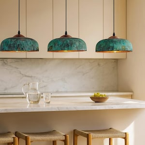 May include: Three pendant lights with verdigris green shades and copper-coloured tops hang above a kitchen counter. The kitchen counter has a clear glass jug, a small glass, and a wooden bowl with green grapes.