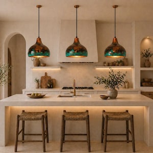 May include: Three copper-colored pendant lights with teal accents hang above a kitchen island with woven stools. The kitchen features a neutral color palette, wooden accents, and a large window with a view of greenery.