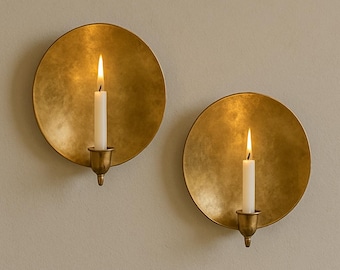 May include: Two wall-mounted candle holders. Each features a round, textured, gold-colored plate with a small brass candle cup. A white candle is lit in each cup, with a bright flame. The candle holders are mounted on a neutral-colored wall.