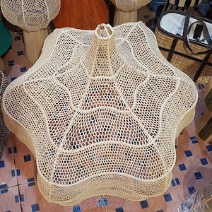 May include: A large, star-shaped woven basket in natural beige tones. The basket has a unique, open-weave design with a central, cylindrical opening. Other woven items are visible in the background, suggesting a home decor theme.