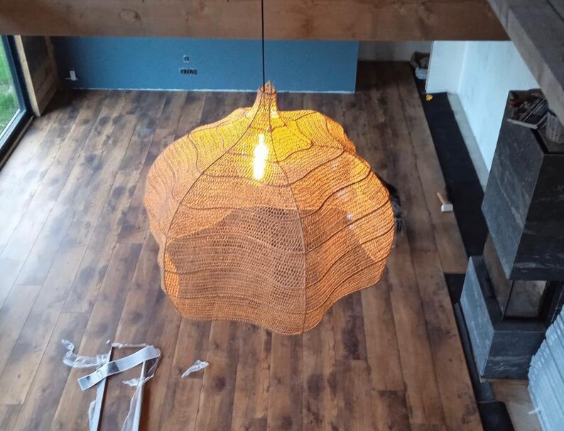 May include: An overhead shot of a woven, spherical pendant light. The light emits a warm orange glow. The fixture hangs above a wooden floor. The room has a modern design with a fireplace and a blue wall.
