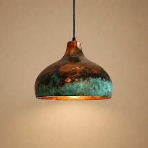 May include: A copper pendant light. The bell-shaped shade has a dark brown top, transitioning to a turquoise and copper finish at the bottom. The light hangs from a black cord. The fixture's diameter is approximately 30 cm.