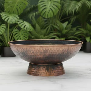 Peut inclure: Un bol en cuivre martelé avec un intérieur bleu foncé repose sur un socle. Le bol, rempli d'eau, est entouré de plantes vertes luxuriantes dans des pots noirs, créant un accent décoratif naturel.