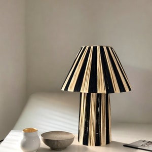 May include: A black and cream striped table lamp with a wide, cylindrical base and a pleated lampshade. The lamp is sitting on a white surface with a small white bowl and a gold-rimmed white cup.