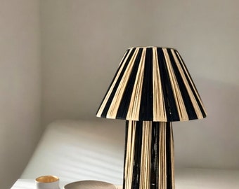 Puede incluir: Una lámpara de mesa decorativa con una pantalla y base a rayas negras y beige. La lámpara es cilíndrica y se encuentra sobre una superficie blanca, junto a un pequeño jarrón blanco con borde dorado y un cuenco texturizado. El diseño de la lámpara es moderno y elegante.