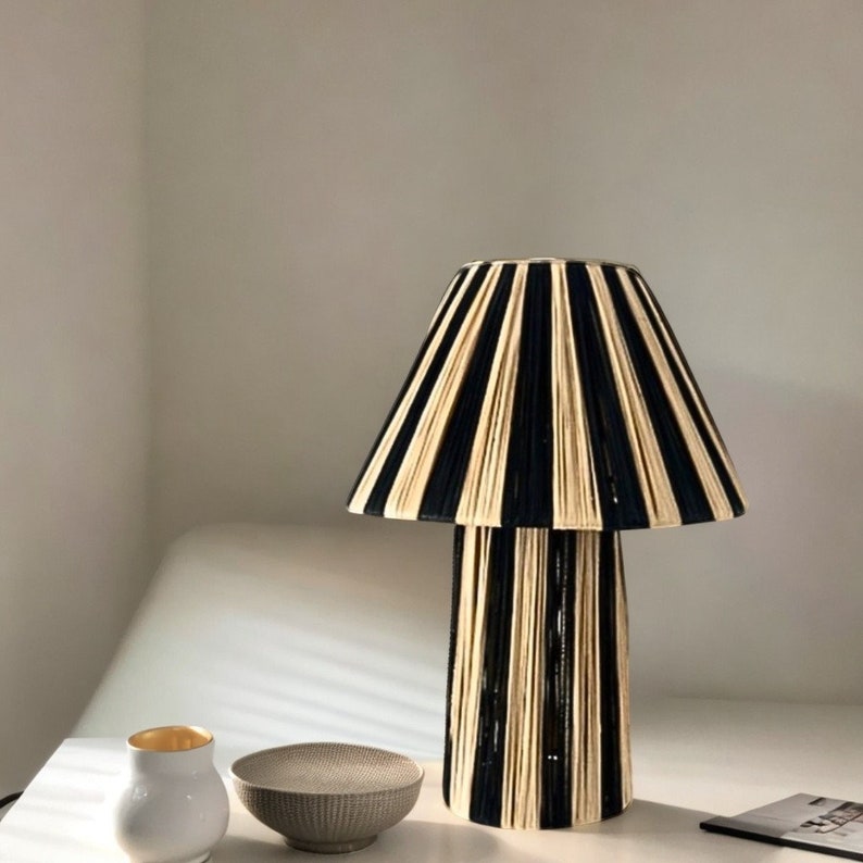 May include: A black and cream striped table lamp with a wide, cylindrical base and a pleated lampshade. The lamp is sitting on a white surface with a small white bowl and a gold-rimmed white cup.