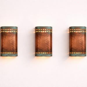 May include: Three copper-toned wall sconces with a rectangular shape. Each sconce features a teal top and bottom with square cut-outs. The sconces are mounted on a white wall and emit a warm light.