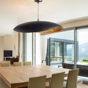 May include: A black and gold pendant light fixture hangs over a wooden dining table with chairs in a modern dining room. The light fixture has a large, round, black shade with a gold interior. The room has large windows with a view of a green lawn and mountains in the distance.