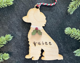 Puede incluir: Un adorno de madera clara con forma de perro que presenta un detalle de hojas de acebo rojas y verdes en el cuello y el nombre "quinn" grabado en letras negras. El adorno cuelga de un cordón trenzado de color rojo, blanco y verde.