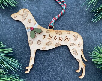 Könnte beinhalten: Ein helles Holzornament in Hundeform mit eingravierten Flecken und dem Namen "oscar" in Schreibschrift. Der Anhänger ist mit einem kleinen grünen Stechpalmenblatt mit roter Beere verziert und besitzt eine rot-weiß-grün gedrehte Aufhängeschnur.