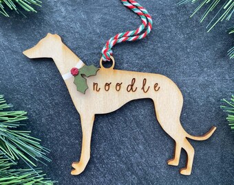 May include: A light-colored wooden ornament shaped like a greyhound dog, featuring the name "noodle" engraved on its side. The dog wears a white collar with green holly leaves and a red berry, and hangs from a red, white, and green twisted cord.