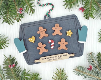 Könnte beinhalten: Ein dunkelgraues Backblech-Ornament aus Holz mit vier Lebkuchenmännchen-Ausschnitten namens Hudson, Hazel, Elise und Gio. Es enthält eine Zuckerstange, zwei gelbe sternförmige Kekse, zwei blaugrüne Ofenhandschuhe und ein Nudelholz mit dem Text GRANDMA'S FAVORITE COOKIES CHRISTMAS 2026.