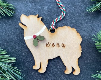 Könnte beinhalten: Ein helles, hundeförmiges Holzornament mit dem eingravierten Namen Xena. Das Ornament verfügt über ein weißes Halsband mit einem kleinen grünen Stechpalmenblatt und einer roten Beere, aufgehängt an einer rot-weiß-grün gedrehten Kordel.