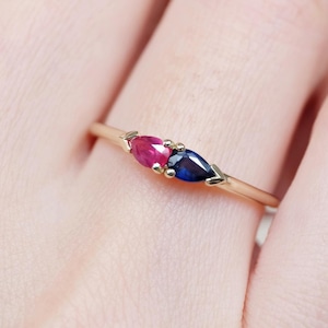 Może przedstawiać: Złoty pierścionek z dwoma kamieniami szlachetnymi w kształcie łezki: żywym czerwonym rubinem i głębokim niebieskim szafirem. Kamienie są umieszczone obok siebie na delikatnej złotej obrączce, tworząc uderzający kontrast kolorów i kształtów. Pierścionek to biżuteria.