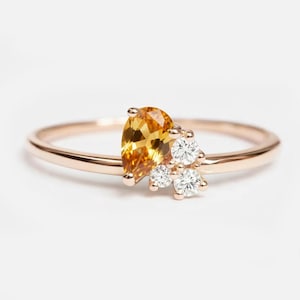 Puede incluir: Un anillo de oro rosa con una piedra preciosa amarilla dorada en forma de pera y tres piedras preciosas redondas y transparentes más pequeñas. El anillo tiene una banda fina y pulida.