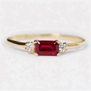 Könnte beinhalten: Ein Goldring mit einem zentralen, smaragdgeschliffenen roten Rubin, der von zwei kleinen runden weißen Diamanten flankiert wird. Der Ring ist schmal und aus poliertem Gold gefertigt, wobei die drei Steine horizontal in sichtbaren Krappen gefasst sind.