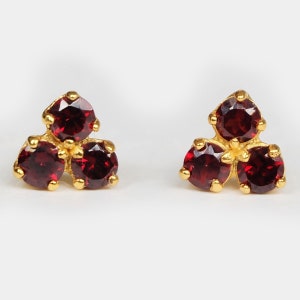 May include: A pair of gold earrings with three red gemstones each. The earrings are small and delicate.