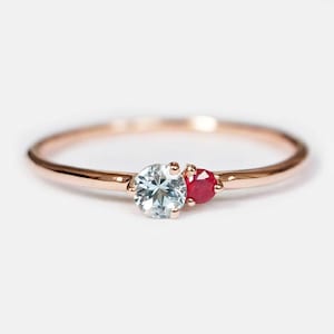 May include: A delicate rose gold ring featuring two round-cut gemstones set side-by-side. The larger stone is a pale blue aquamarine, while the smaller stone is a vibrant red ruby. Both stones are secured in a simple prong setting on a thin band.