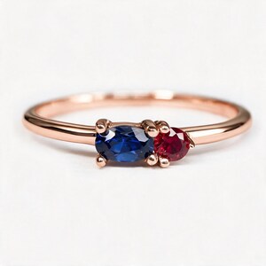 Pode incluir: Um anel de ouro rosa com uma safira azul de corte oval horizontal e um rubi vermelho menor de corte redondo colocados lado a lado. A banda é fina e polida, apresentando uma montagem de metal simples que segura ambas as pedras preciosas.