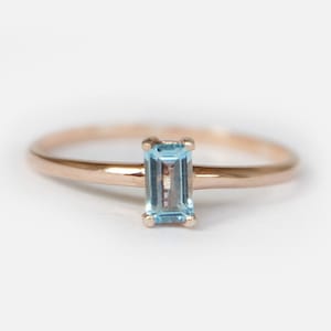May include: A delicate rose gold ring featuring a rectangular, light blue gemstone. The gemstone is set in a four-prong setting, and the band is thin and polished. The ring is displayed against a white background.