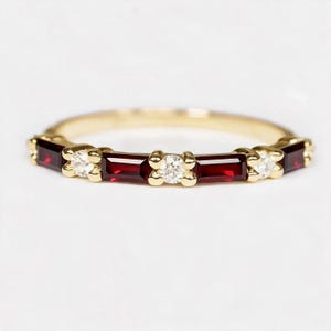 May include: A gold band ring featuring an alternating pattern of four deep red baguette-cut garnets and three clear round-cut diamonds set in a prong setting, creating a continuous horizontal design across the front of the jewelry piece.