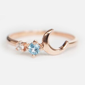 May include: A rose gold ring with a crescent moon design and two gemstones. The larger gemstone is a light blue topaz and the smaller gemstone is a clear diamond.