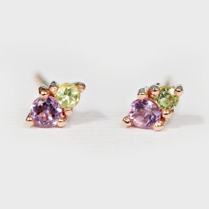 Puede incluir: Un par de pequeños pendientes de botón con tres piedras preciosas. Los pendientes tienen una piedra preciosa rosa en el centro, rodeada de dos piedras preciosas amarillo-verdosas más pequeñas.