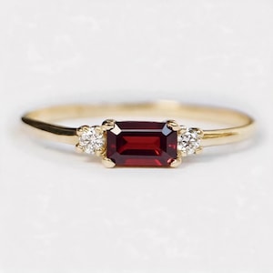Op de afbeelding: Een gouden ring met een centrale smaragdgeslepen dieprode granaatsteen, geflankeerd door twee kleine ronde briljantgeslepen heldere diamanten. De ringband is dun en gepolijst en toont een klassiek ontwerp met drie stenen in klauwzettingen.