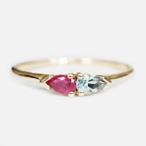 May include: A gold ring with two pear-cut gemstones. One is a bright magenta, the other a pale blue. The ring has a slender, polished band, with the gemstones set closely together.