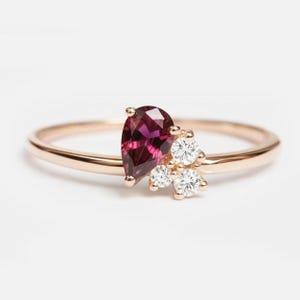 Puede incluir: Un anillo de oro rosa con una piedra preciosa roja oscura en forma de pera y tres piedras preciosas redondas y transparentes más pequeñas. El anillo tiene una banda delicada y un diseño de racimo.