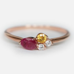 Op de afbeelding: Een ring van roségoud met een rode peervormige edelsteen, een gele ronde edelsteen en drie kleinere witte ronde edelstenen.