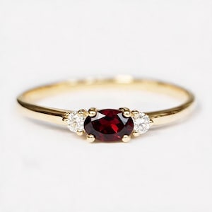 May include: A gold ring featuring a central oval-cut deep red garnet gemstone flanked by two small round-cut clear white diamonds. The band is smooth and polished, showcasing a classic three-stone design with prong settings for each stone.