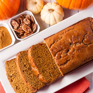 Op de afbeelding: Een snee pumpkinbrood op een witte schaal met een rode servet. Er zijn ook twee kleine kommen met pecannoten en kaneel op de schaal. Er zijn ook twee hele pompoenen en twee kleine pompoenen op de tafel.