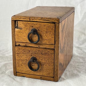 May include: A small wooden chest with two stacked drawers, each featuring a dark metal ring pull handle. The exterior displays a natural wood grain pattern in shades of light and medium brown, with a smooth top surface and rectangular frame.
