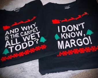 May include: Two black t-shirts with a Christmas theme. The first shirt says "And why is the carpet all wet Todd?" The second shirt says "I don't know, Margo!"