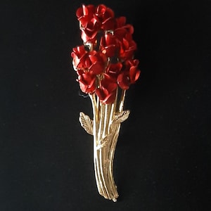 May include: A vintage brooch featuring a bouquet of red roses. The roses are clustered together, with a gold-tone stem and leaves. The brooch is likely made of metal and is a classic floral design.